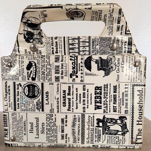 May include: Vintage-style metal carrier with a handle, covered in a black and white newspaper print. The carrier has a rectangular shape with rounded corners and is secured with silver rivets. The print includes advertisements and illustrations.