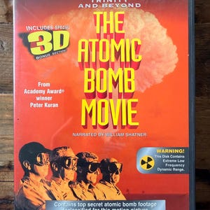 May include: DVD case for "The Atomic Bomb Movie" with a red and yellow design. The cover features a mushroom cloud graphic and the text "The Atomic Bomb Movie." Includes "3D Bonus Section" and "Digital Collector's Edition."