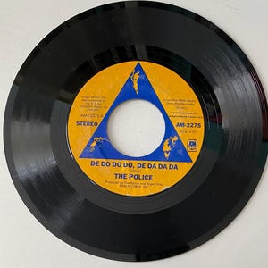 May include: A black vinyl record with a yellow and blue label. The label features a triangle with a world map inside. The title of the record is "De Do Do Do, De Da Da Da" by The Police. The record is from the A&M album "Zenyatta Mondatta" and has a time of 4:09.