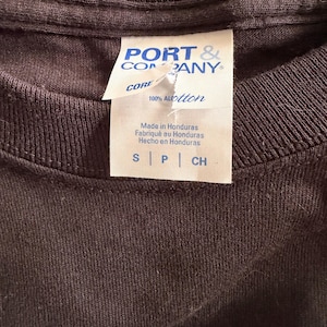 May include: Close-up of a dark brown t-shirt collar with a white tag. The tag reads "PORT & COMPANY CORE" and "100% ALOtton". The tag also states "Made in Honduras" in English, French, and Spanish. The shirt size is indicated as S, P, and CH.