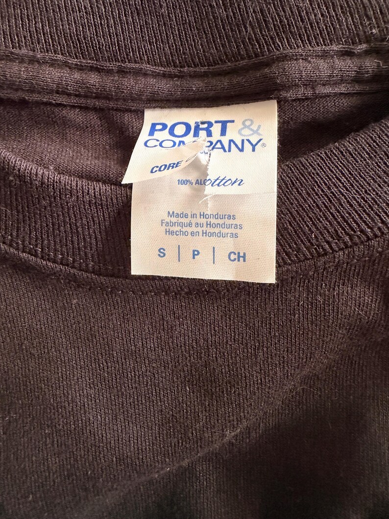 May include: Close-up of a dark brown t-shirt collar with a white tag. The tag reads "PORT & COMPANY CORE" and "100% ALOtton". The tag also states "Made in Honduras" in English, French, and Spanish. The shirt size is indicated as S, P, and CH.
