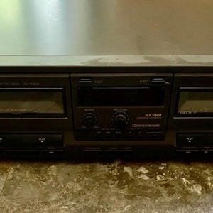 May include: A black Technics RS-TR373 stereo cassette deck. The deck has two cassette slots, a central control panel with buttons and a display, and the Technics logo on the top left.