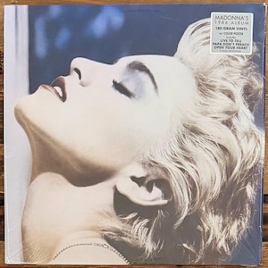 May include: A square vinyl record cover featuring a sepia-toned portrait of Madonna with her head tilted back. A white sticker in the top right corner reads: MADONNA'S 1986 ALBUM, 180 GRAM VINYL, w/ COLOR POSTER, Includes LIVE TO TELL, PAPA DON'T PREACH, OPEN YOUR HEART.