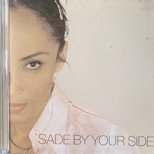 May include: A close-up of a woman's face, partially obscured by a CD case. She is wearing a light pink shirt and has dark hair. The text "SADE BY YOUR SIDE" is printed in red on the CD case.