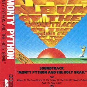 May include: A red cassette tape case with the title "Monty Python and the Holy Grail" in white text. The case features a sunset scene with a yellow and orange sky and green grass. The text "Soundtrack" is printed in white on a red background. The text "Executive Version" is printed in white on a black background.