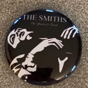 May include: Black and white button featuring a silhouette of a person with the text "THE SMITHS The Queen Is Dead" printed on the button.