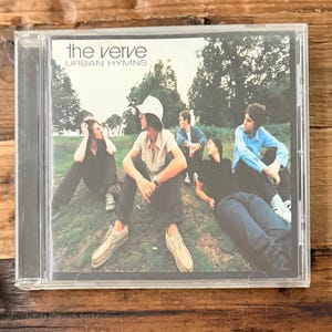 May include: A clear plastic CD jewel case containing the album Urban Hymns by The Verve. The cover art features four individuals sitting on a grassy hill, with one person wearing a white bucket hat, light shirt, and tan shoes, and others in casual attire.