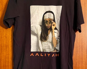 May include: Black t-shirt featuring a portrait of Aaliyah wearing sunglasses and a bandana. The image is in a rectangular frame, with the name "AALIYAH" printed in orange below the portrait. The shirt is hanging on a wooden hanger.