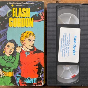 May include: A vintage VHS tape and its box for "Flash Gordon." The box features a comic-style illustration of a man and woman in space suits. The tape is black with white labels.