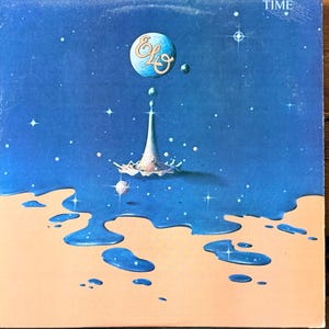 May include: Album cover art for "Time" by ELO. The artwork features a blue and tan landscape with a stylized water droplet and a floating sphere with the band's logo. The word "TIME" is at the top.