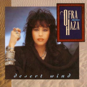 May include: A woman with long dark hair and dark eyes is wearing a black shirt and silver jewelry. The album cover features the text "Ofra Haza" and "Desert Wind".