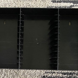 May include: Black plastic VHS tape storage case with three compartments. The case has a label that reads "Case Logic".