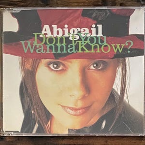 May include: CD case featuring a woman with long hair and a hat. The text "Abigail Don't You Wanna Know?" is displayed in white and green. The woman is wearing a black jacket.