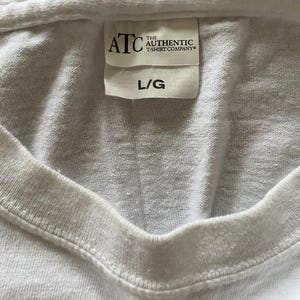 Puede incluir: Primer plano de una camiseta blanca con cuello en V. La etiqueta dice "ATC THE AUTHENTIC T-SHIRT COMPANY" y "L/G". La tela parece ser una mezcla de algod&oacute;n suave. La camiseta es probablemente una prenda b&aacute;sica.