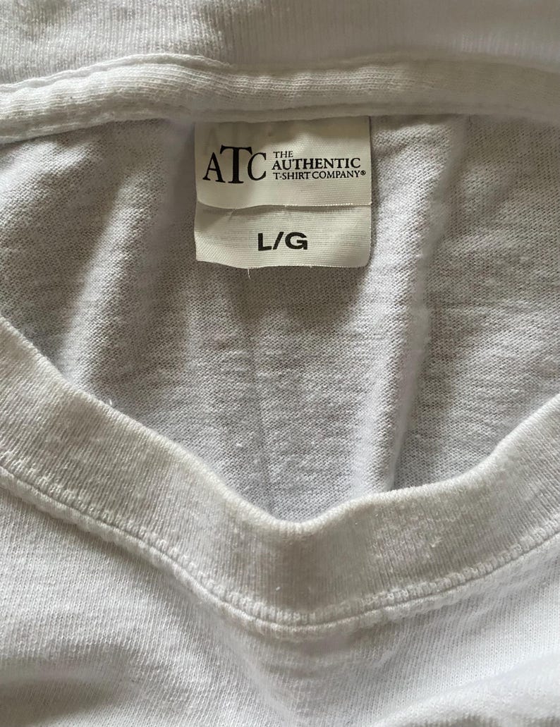 Puede incluir: Primer plano de una camiseta blanca con cuello en V. La etiqueta dice "ATC THE AUTHENTIC T-SHIRT COMPANY" y "L/G". La tela parece ser una mezcla de algod&oacute;n suave. La camiseta es probablemente una prenda b&aacute;sica.