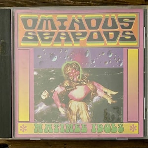 May include: A CD case with a colorful album cover. The album art features the text "OMINOUS SEAPODS" in a retro font above an illustration of a figure holding another figure in a cosmic setting. The text "MATINEE IDOLS" is below the illustration.