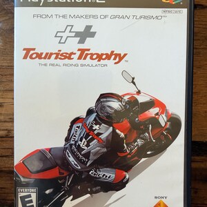 May include: A PlayStation 2 game case for "Tourist Trophy: The Real Riding Simulator." The cover features a red and black motorcycle with a rider in racing gear. The game's title is in red, with the PlayStation 2 logo at the top.