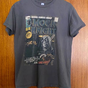 May include: Gray t-shirt featuring a comic book cover design. The design includes the text "MOON KNIGHT" and "MARVEL COMICS GROUP". The shirt also has the text "I CAN'T TELL THE DIFFERENCE BETWEEN MY WAKING LIFE AND DREAMS!"