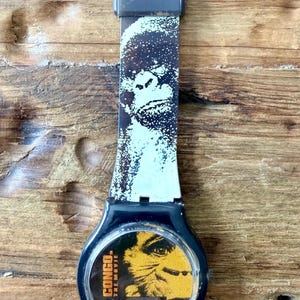 May include: Black wristwatch featuring a design from the movie "Congo." The watch band has a white and black pattern with a gorilla image. The watch face displays a yellow and black image of a gorilla and the movie title.