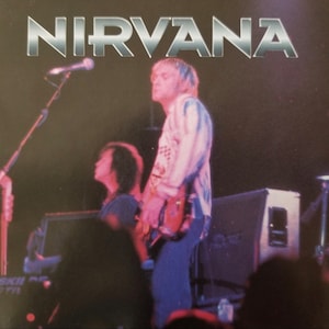 May include: A black and white photo of a band performing on stage. The band is called "NIRVANA". The lead singer is wearing a white and blue patterned shirt and is playing a guitar. The other band member is wearing a black shirt and is playing a bass guitar.