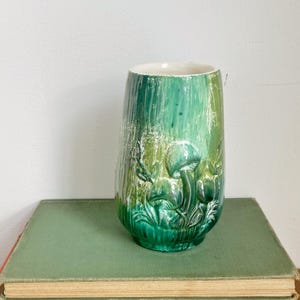 May include: A green ceramic vase with a textured surface and a mushroom design. The vase is cylindrical with a wide base and a narrower opening. The color gradient ranges from dark to light green.