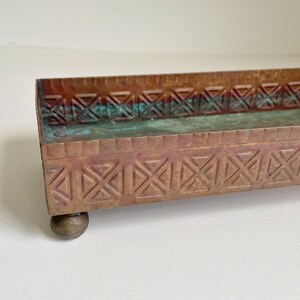 May include: A rectangular copper tray featuring a repeating embossed geometric cross pattern along the sides. The metal surface displays a weathered patina with visible green oxidation. The tray is supported by small, rounded metal feet.