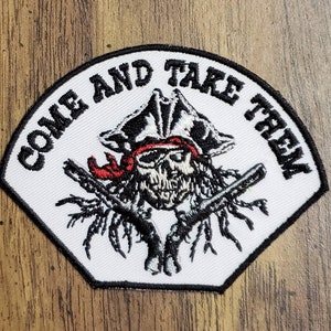 Puede incluir: Parche blanco bordado con una calavera con un sombrero de pirata y sosteniendo dos pistolas. El texto "COME AND TAKE THEM" está escrito alrededor de la calavera.