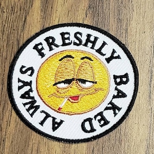 May include: A round, white patch with black stitching and the text "ALWAYS FRESHLY BAKED" around a yellow cartoon face with a cigarette in its mouth.