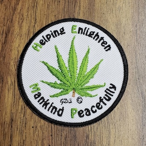 May include: Round embroidered patch with a cannabis leaf in the center. The patch has a white background with the words "Helping Enlighten Mankind Peacefully" in green and black. The patch is outlined in black.