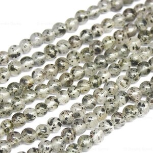 May include: A strand of round, translucent, light grey beads with black flecks. The beads are strung together on a white background.
