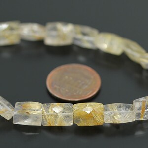May include: A strand of golden rutilated quartz beads, each bead is a square shape with a faceted cut. The beads are strung on a white cord and are displayed on a black surface.