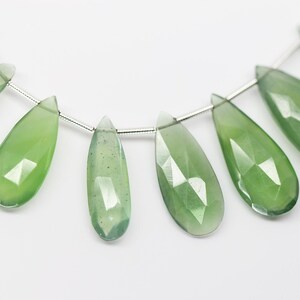 May include: A strand of green gemstone beads, each bead is faceted and shaped like a teardrop. The beads are strung on a silver wire.