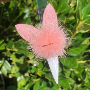 May include: A pink fuzzy bunny charm with a white nail tip and a clear rhinestone. The charm is attached to a clear plastic stick.