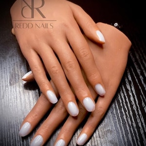 May include: Close-up of a pair of hands with white oval-shaped nails. The nails are painted a milky white colour. The hands are resting on a wooden surface. The text "REDD NAILS" is visible in the top left corner of the image.