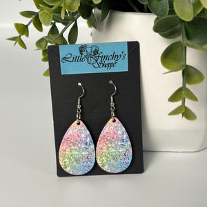 May include: A pair of teardrop-shaped earrings featuring a vibrant rainbow-coloured glitter pattern with a subtle leopard print overlay. The earrings hang from silver-toned metal hooks and are displayed on a black card labelled Little Finchy's Shoppe.
