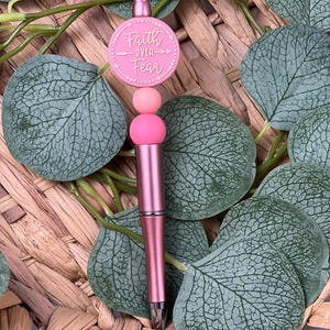 Könnte beinhalten: Ein rosafarbener Stift mit den Worten "Faith Over Fear" auf einem runden Anhänger. Der Stift hat eine metallische Oberfläche und ist mit rosa Perlen verziert. Der Stift befindet sich vor einem Hintergrund aus grünen Blättern und geflochtenem Korbmaterial.