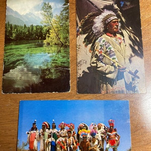 May include: Three vintage postcards featuring Native American imagery. The first postcard shows a lake with mountains in the background. The second postcard shows a Native American man wearing a headdress and holding a pipe. The third postcard shows a group of Native Americans in traditional clothing.