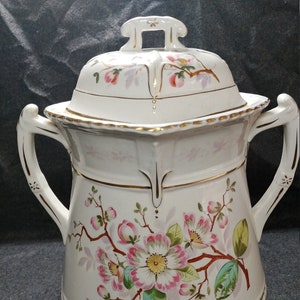 May include: White ceramic jar with a lid and two handles. The jar has a floral design with pink and green flowers. The lid has a gold trim and a small handle.