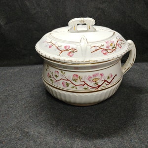 May include: White ceramic chamber pot with a lid, decorated with pink flowers and gold accents. The pot has a handle and a floral design.