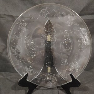 May include: A clear glass plate with an etched design of a snowman, a person in a long coat, and another person in a hat. The plate is surrounded by a circle of smaller etched designs, including snowflakes, ornaments, and presents.