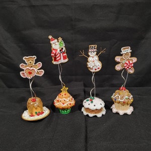 May include: Five Christmas-themed gingerbread ornaments, including a snowman, a gingerbread man, a teddy bear, a cupcake with a star on top, and a cupcake with a holly sprig on top.