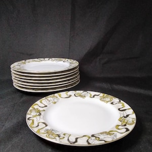 May include: A stack of ten white ceramic plates with a black and gold design featuring cherubs and ribbons. The plates are stacked on a black surface.