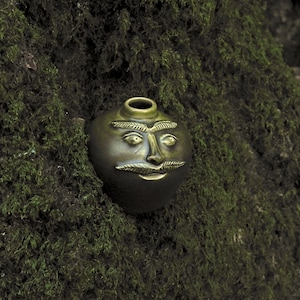 May include: A bronze-coloured, round ceramic vessel with a face design, featuring a moustache, eyebrows, and eyes. The vessel has a small opening at the top, set against green moss.
