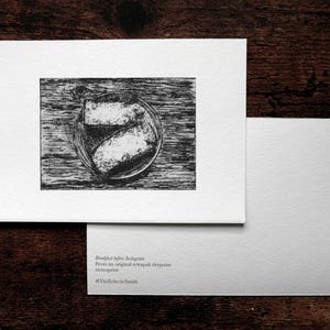 May include: A monochrome print of a breakfast scene on a white card, set against a dark wooden background. A second blank card is next to it. The text on the card reads "Breakfast before Instagram" and "#TheEclecticSmith".