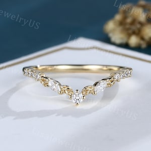 Puede incluir: Un anillo dorado en forma de V con un diamante redondo central y piedras más pequeñas en forma de marquesa. El anillo está engastado con pequeñas piedras transparentes a lo largo de la banda. El anillo está sobre una superficie blanca con un fondo azul oscuro.