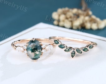 Peut inclure: Un ensemble de deux bagues en or rose avec de l'agate mousse verte et des diamants blancs. La bague de fiançailles présente une grande agate mousse ovale entourée de diamants plus petits. La bague de mariage est une bande délicate avec une rangée de pierres d'agate mousse verte.