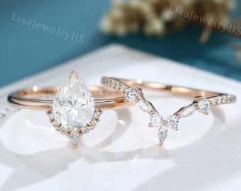 May include: A pear-shaped diamond engagement ring set in rose gold with a delicate, v-shaped wedding band. The engagement ring features a halo of smaller diamonds around the center stone and the wedding band is adorned with small diamonds.