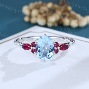 May include: A silver ring featuring an oval, light blue gemstone, flanked by smaller, red gemstones. The band has a braided design. The ring is set against a white surface with a blurred background.
