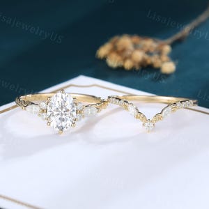 May include: A gold engagement ring set featuring a central oval-cut stone flanked by marquise-cut stones on a textured band, paired with a matching V-shaped chevron wedding band adorned with small marquise and round-cut stones in a leaf pattern.