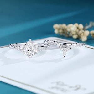 May include: A silver-toned ring set featuring a pear-cut center stone flanked by smaller marquise and round stones on a twisted band, paired with a matching V-shaped chevron band adorned with a series of small round and marquise-cut stones.